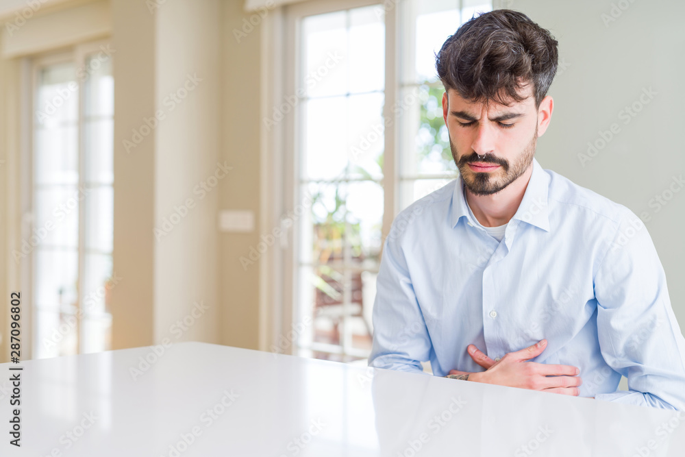 Young businesss man sitting on white table with hand on stomach because nausea, painful disease feeling unwell. Ache concept. Young businesss man sitting on white table with hand on stomach because nausea, painful disease feeling unwell. Ache concept.