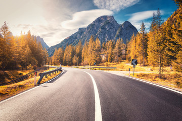 Road in autumn forest at sunset in Italy. Beautiful mountain roadway, trees with orange foliage and sunlight. Landscape with empty asphalt road through woodland, blue sky, high rocks in fall. Travel