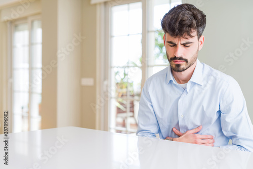 Young businesss man sitting on white table with hand on stomach because nausea, painful disease feeling unwell. Ache concept. © Krakenimages.com