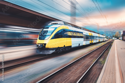Fototapeta Wysoki prędkość żółty pociąg w ruchu na stacji kolejowej o zachodzie słońca. Nowoczesny miejski pociąg pasażerski z efektem rozmycia ruchu na peronie kolejowym. Przemysłowy. Tło kolejowe i niewyraźne