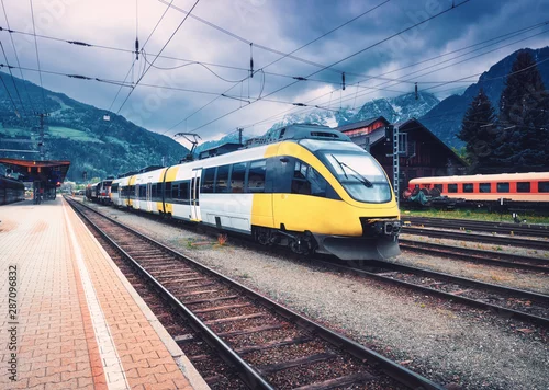 Naklejka na ścianę Piękny pociąg dużych prędkości na stacji kolejowej w górach o zachodzie słońca jesienią Żółty nowoczesny pociąg podmiejski na peronie kolejowym. Krajobraz przemysłowy z kolei. Transport pasażerski