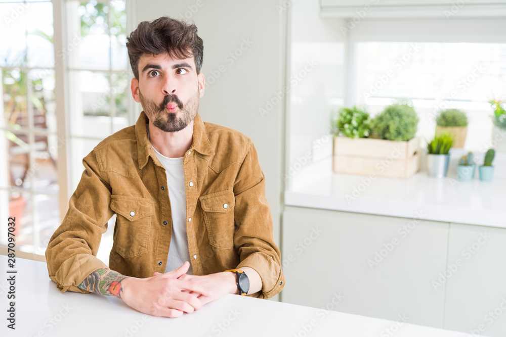 Young man wearing casual jacket sitting on white table making fish face with lips, crazy and comical gesture. Funny expression. Young man wearing casual jacket sitting on white table making fish face with lips, crazy and comical gesture. Funny expression.