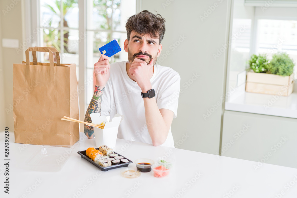 Young man eating asian sushi from home delivery using credit card as payment serious face thinking about question, very confused idea Young man eating asian sushi from home delivery using credit card as payment serious face thinking about question, very confused idea