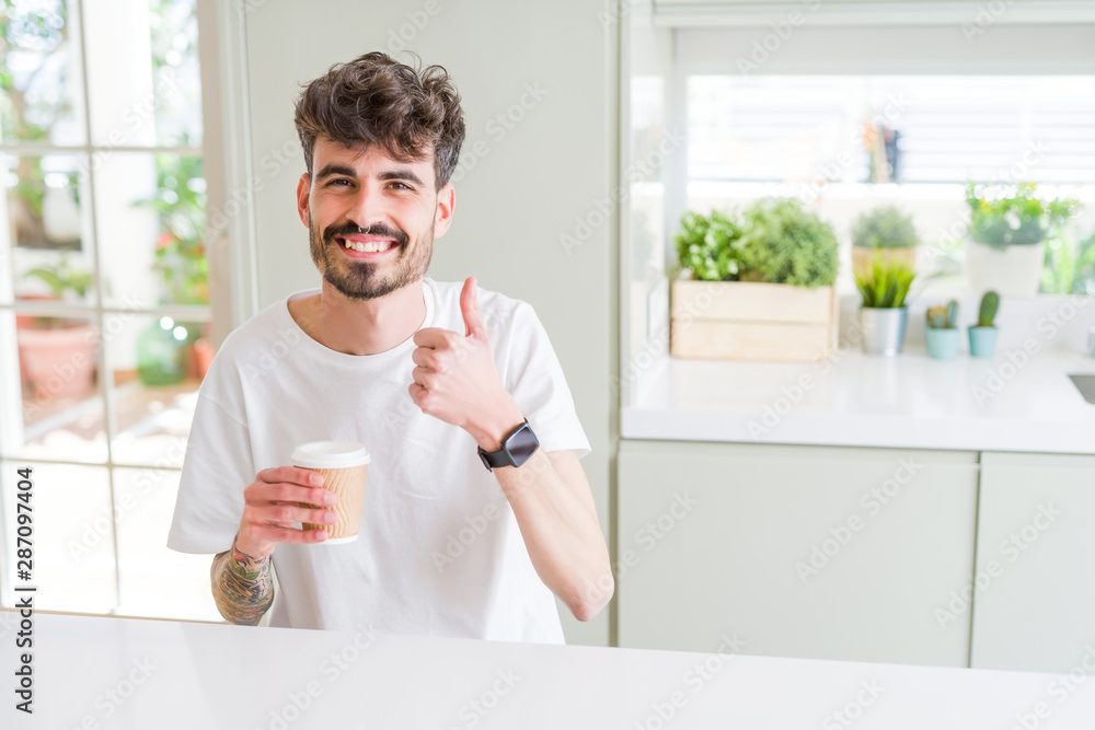 Young man drinking a coffee in a paper cup in the morning happy with big smile doing ok sign, thumb up with fingers, excellent sign Young man drinking a coffee in a paper cup in the morning happy with big smile doing ok sign, thumb up with fingers, excellent sign