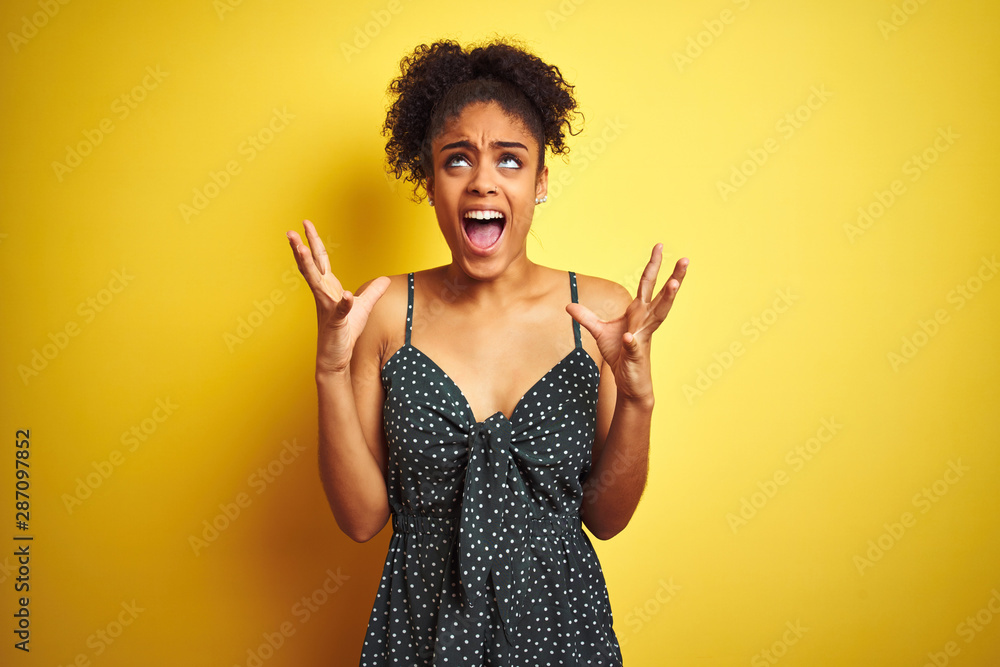 African american woman wearing summer casual green dress over isolated yellow background crazy and mad shouting and yelling with aggressive expression and arms raised. Frustration concept. African american woman wearing summer casual green dress over isolated yellow background crazy and mad shouting and yelling with aggressive expression and arms raised. Frustration concept.