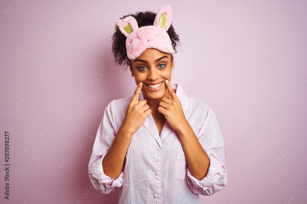 Young african american woman wearing pajama and mask over isolated pink background Smiling with open mouth, fingers pointing and forcing cheerful smile Young african american woman wearing pajama and mask over isolated pink background Smiling with open mouth, fingers pointing and forcing cheerful smile
