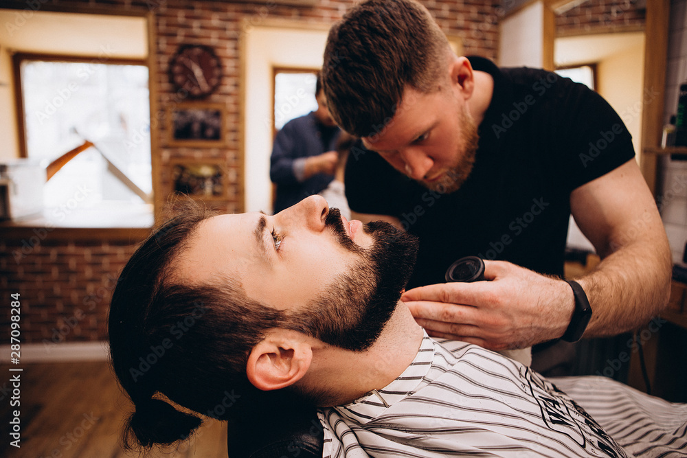 Getting perfect shape. Close-up side view of young stylish bearded man getting beard haircut by bearded, muscular hairdresser or barber at barbershop Getting perfect shape. Close-up side view of young stylish bearded man getting beard haircut by bearded, muscular hairdresser or barber at barbershop