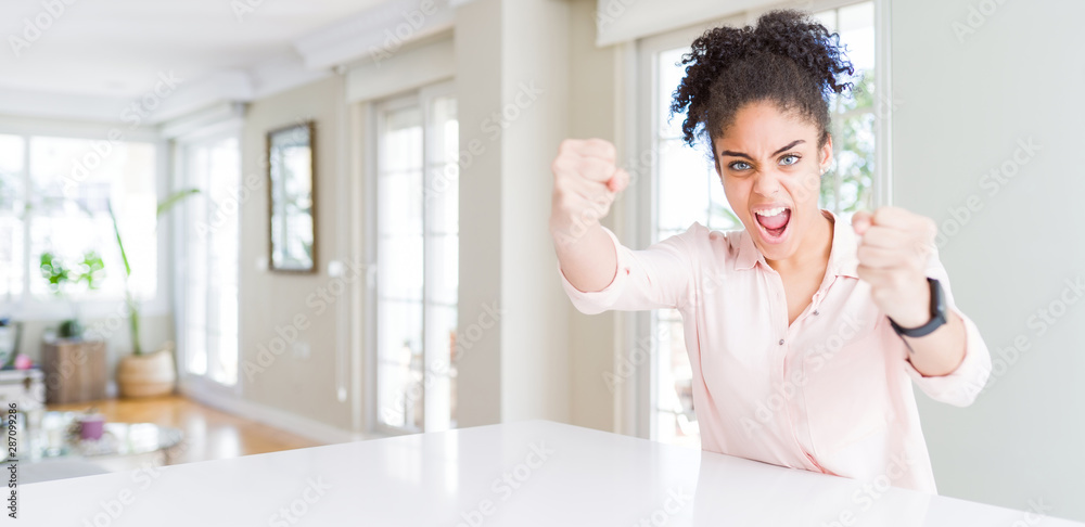 Wide angle of beautiful african american woman with afro hair angry and mad raising fists frustrated and furious while shouting with anger. Rage and aggressive concept.