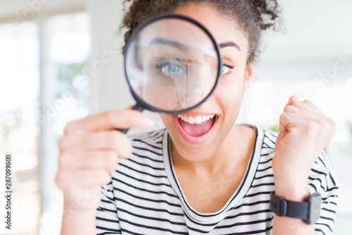 Young african american woman looking through magnifying glass screaming proud and celebrating victory and success very excited, cheering emotion