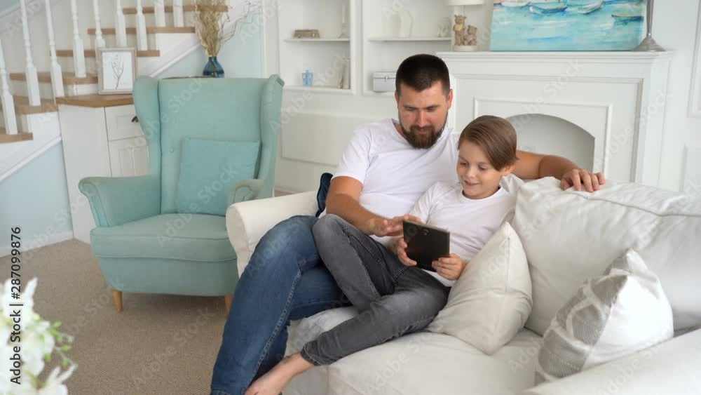 Father and son game players fans sit together at home on sofa, using the tablet. Father and son game players fans sit together at home on sofa, using the tablet.