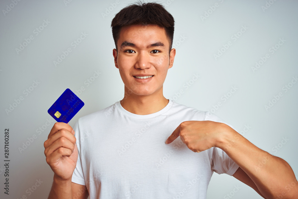 Young asian chinese man holding credit card standing over isolated white background with surprise face pointing finger to himself Young asian chinese man holding credit card standing over isolated white background with surprise face pointing finger to himself