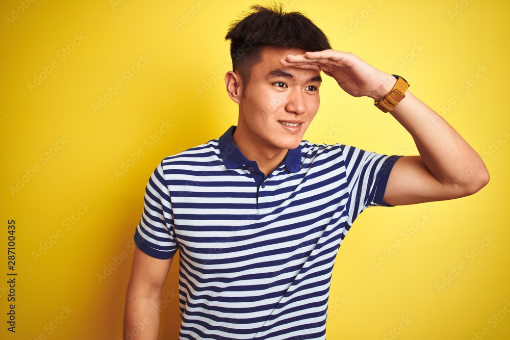 Young asian chinese man wearing striped polo standing over isolated yellow background very happy and smiling looking far away with hand over head. Searching concept. Young asian chinese man wearing striped polo standing over isolated yellow background very happy and smiling looking far away with hand over head. Searching concept.