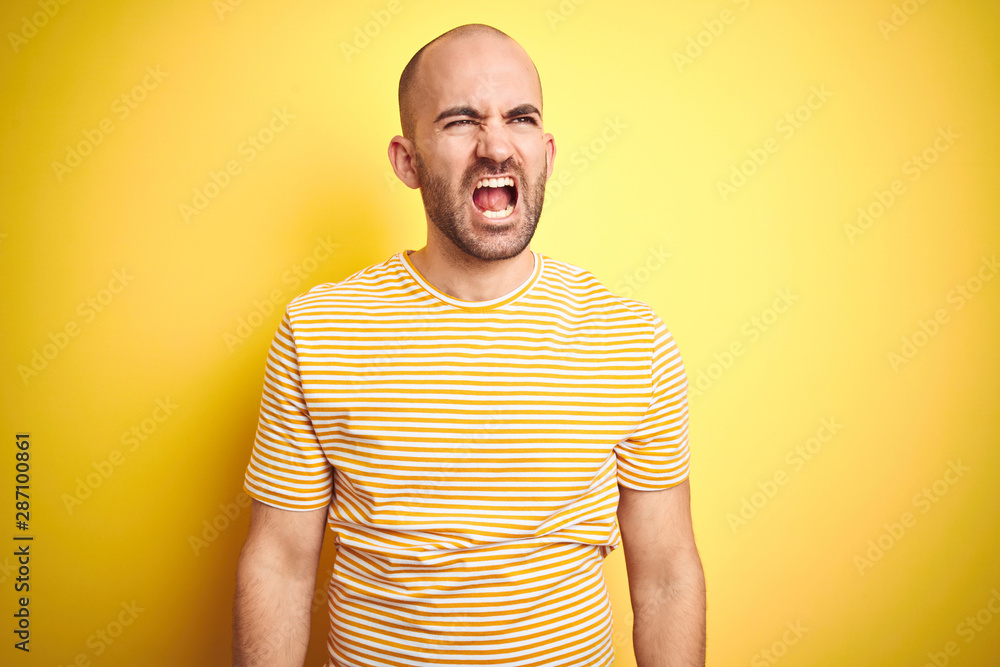 Young bald man with beard wearing casual striped t-shirt over yellow isolated background angry and mad screaming frustrated and furious, shouting with anger. Rage and aggressive concept. Young bald man with beard wearing casual striped t-shirt over yellow isolated background angry and mad screaming frustrated and furious, shouting with anger. Rage and aggressive concept.