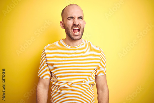 Young bald man with beard wearing casual striped t-shirt over yellow isolated background angry and mad screaming frustrated and furious, shouting with anger. Rage and aggressive concept.