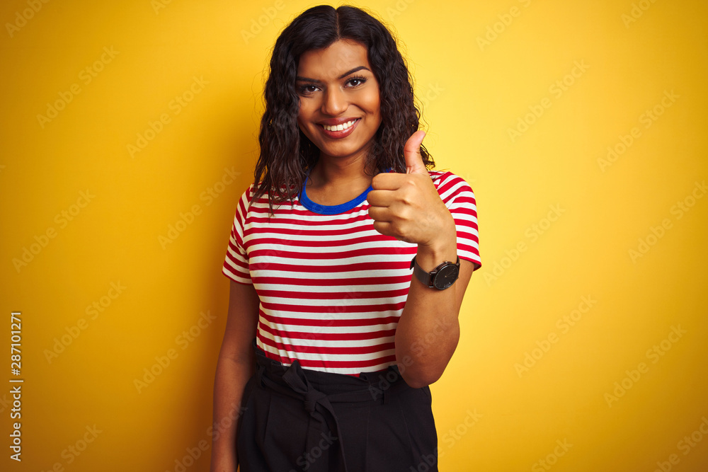 Transsexual transgender woman wearing stiped t-shirt over isolated yellow background doing happy thumbs up gesture with hand. Approving expression looking at the camera with showing success. Transsexual transgender woman wearing stiped t-shirt over isolated yellow background doing happy thumbs up gesture with hand. Approving expression looking at the camera with showing success.