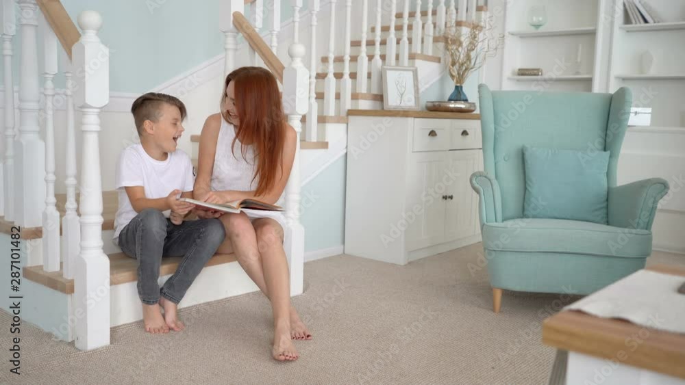 son and mom sits on the stairs and read a book . They look at camera and smile son and mom sits on the stairs and read a book . They look at camera and smile
