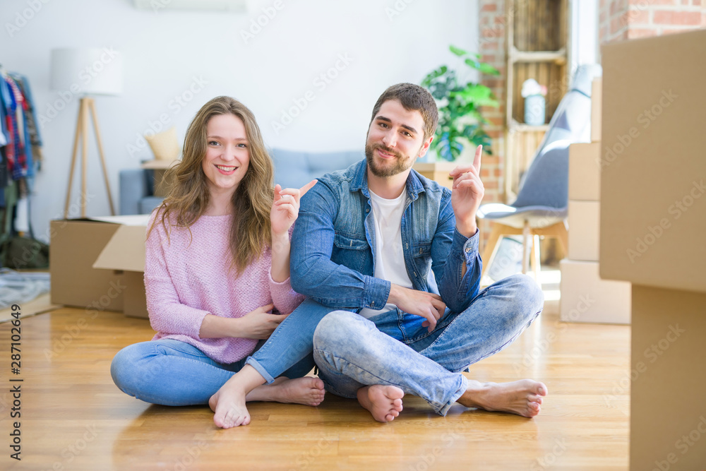 Young beautiful couple moving to a new house sitting on the floor with a big smile on face, pointing with hand and finger to the side looking at the camera. Young beautiful couple moving to a new house sitting on the floor with a big smile on face, pointing with hand and finger to the side looking at the camera.