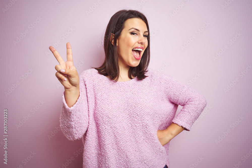 Young beautiful brunette woman wearing a sweater over pink isolated background smiling with happy face winking at the camera doing victory sign. Number two. Young beautiful brunette woman wearing a sweater over pink isolated background smiling with happy face winking at the camera doing victory sign. Number two.