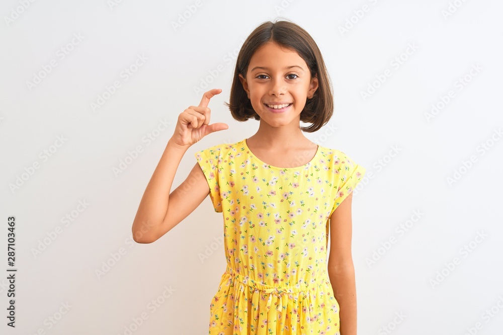 Young beautiful child girl wearing yellow floral dress standing over isolated white background smiling and confident gesturing with hand doing small size sign with fingers looking and the camera Young beautiful child girl wearing yellow floral dress standing over isolated white background smiling and confident gesturing with hand doing small size sign with fingers looking and the camera