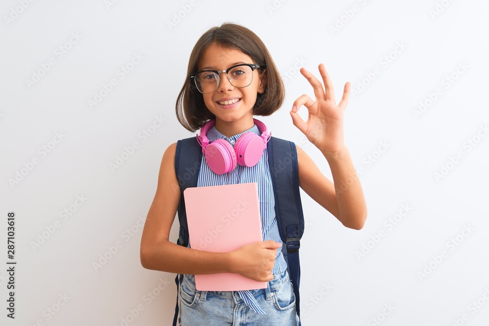 Student child girl wearing backpack glasses book headphones over isolated white background doing ok sign with fingers, excellent symbol Student child girl wearing backpack glasses book headphones over isolated white background doing ok sign with fingers, excellent symbol