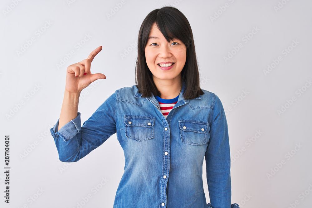 Young beautiful chinese woman wearing denim shirt over isolated white background smiling and confident gesturing with hand doing small size sign with fingers looking and the camera. Measure concept. Young beautiful chinese woman wearing denim shirt over isolated white background smiling and confident gesturing with hand doing small size sign with fingers looking and the camera. Measure concept.