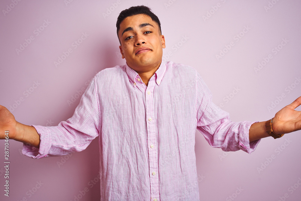 Young brazilian man wearing shirt standing over isolated pink background clueless and confused expression with arms and hands raised. Doubt concept. Young brazilian man wearing shirt standing over isolated pink background clueless and confused expression with arms and hands raised. Doubt concept.