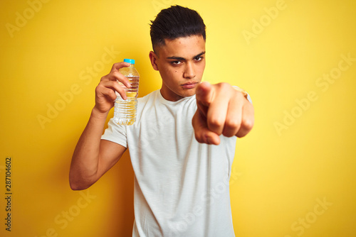 Young brazilian man holding bottle of water standing over isolated yellow background pointing with finger to the camera and to you, hand sign, positive and confident gesture from the front © Krakenimages.com