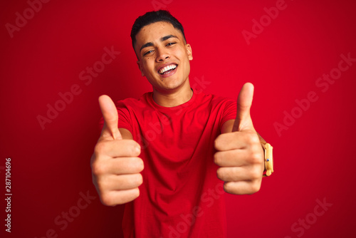 Young brazilian man wearing t-shirt standing over isolated red background success sign doing positive gesture with hand, thumbs up smiling and happy. Cheerful expression and winner gesture. © Krakenimages.com