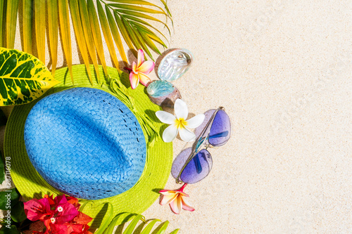 Blue and green straw hat with sunglasses, sea shells and frangipani flowers with green palm leafs on sand, summer vacation concept, top view, copy sapce