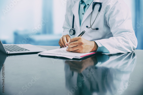 Male doctor sitting at table and writing on a document report in hospital office. Medical healthcare staff and doctor service. photo
