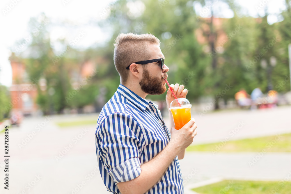 Fototapeta premium Portrait of a positive cheerful young man with a glass of juice with a straw while walking in the park on a warm sunny summer day. The concept of rest after study and work on weekends.