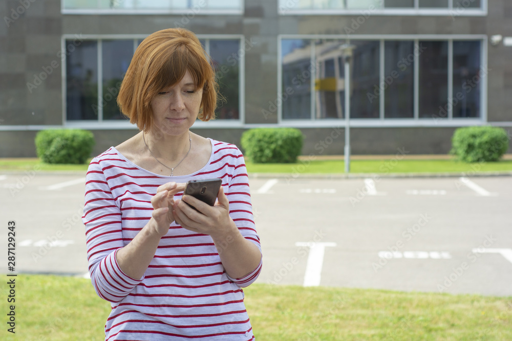 Red-haired middle-aged woman with freckles thoughtfully typing a message on the screen of a mobile phone Red-haired middle-aged woman with freckles thoughtfully typing a message on the screen of a mobile phone