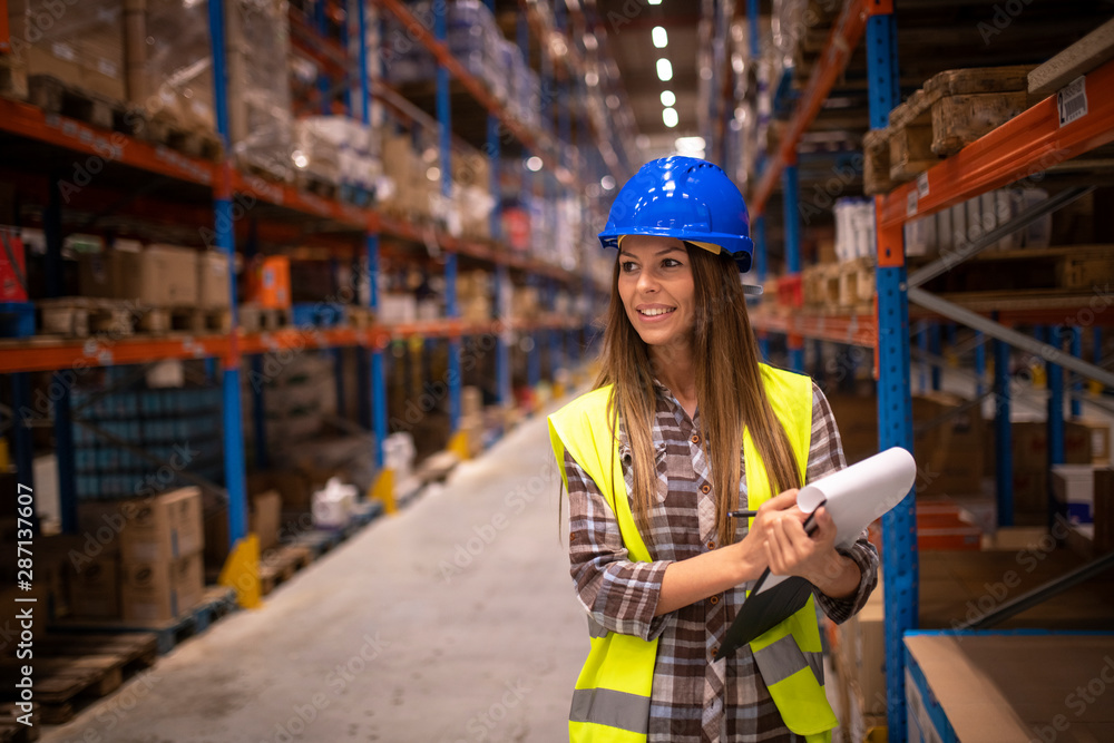 Portrait of cheerful woman in protective uniform checking packages and stock of products in warehouse storage room. Industrial worker controlling distribution in storehouse. Portrait of cheerful woman in protective uniform checking packages and stock of products in warehouse storage room. Industrial worker controlling distribution in storehouse.