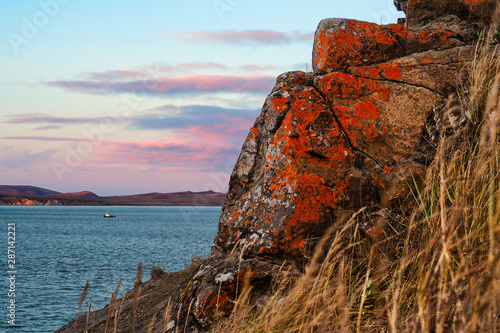 Evening seascape. The coast of the Anadyr estuary. Rocks are covered with lichen. A boat is floating in the distance. Cape Alexander, Chukotka, Siberia, Far East Russia. Arctic landscapes and nature. © Andrei Stepanov