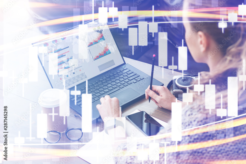 Candlestick chart in foreground. In background woman working on laptop computer with graphs, charts, diagrams on monitor.