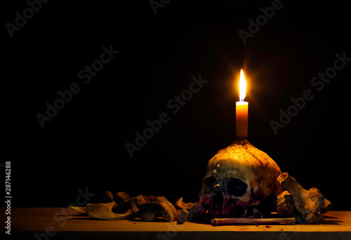 Skull which has candle on head and lighting from candle and pile of bone on wooden plank in dark light room