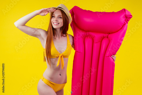 Redhaired ginger female posing floating with inflatable mattress wearing swimmingsuit and hat, looking away at copy space in studio yellow background.Where is water? © yurakrasil