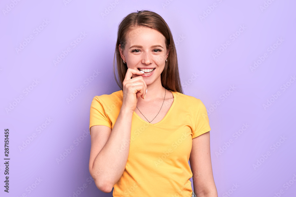 Front view of pretty playful girl with intriguing face expression, keeps hand near lips, thinking about something pleasant and funny, looking in camera with cunning smile, isolated over purple wall. Front view of pretty playful girl with intriguing face expression, keeps hand near lips, thinking about something pleasant and funny, looking in camera with cunning smile, isolated over purple wall.