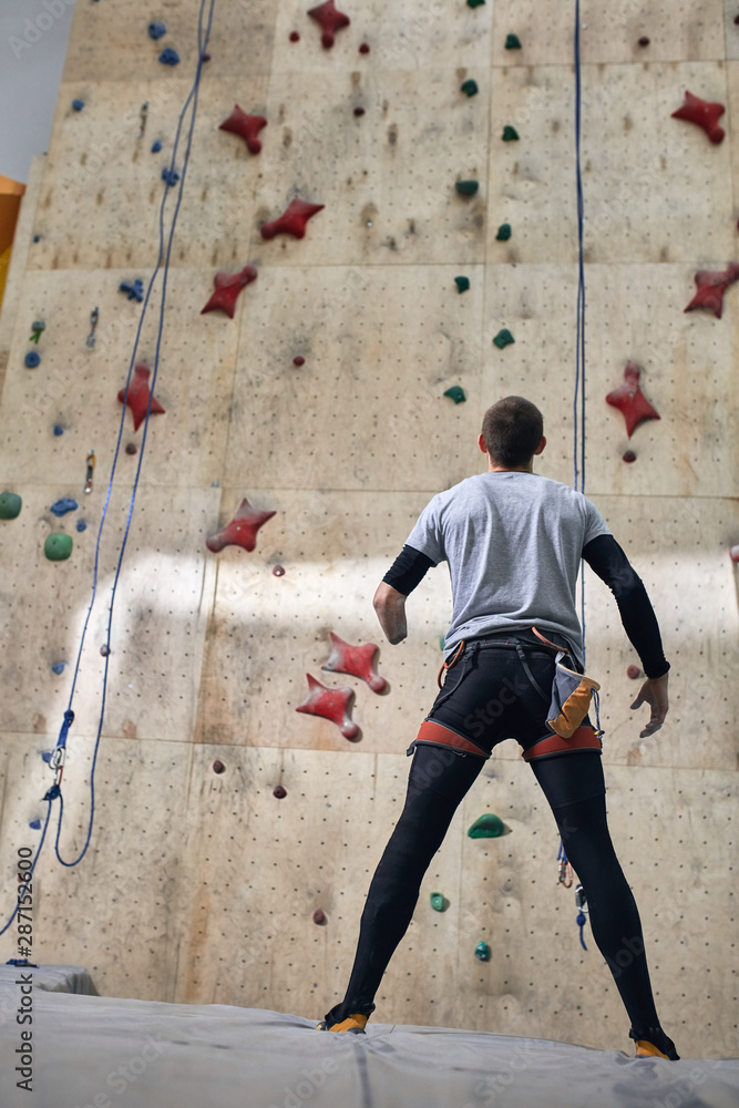 Full length back view of strong motivated rock-climber without forearm,  standing in front of high artificial climbing wall, a wooden panel with  holds and ropes, ready to start bouldering training. Photos