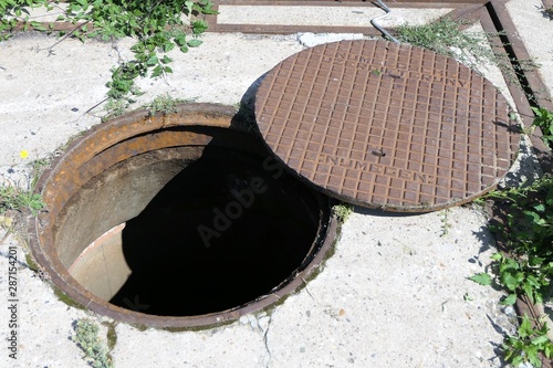Old rusty open manhole surrounded by weeds