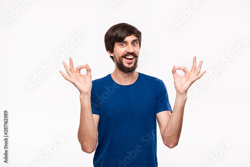 Handsome brunet bearded man with mustaches in a blue shirt showing gesture of OK sign with two hands standing isolated over white background. © ianachyrva