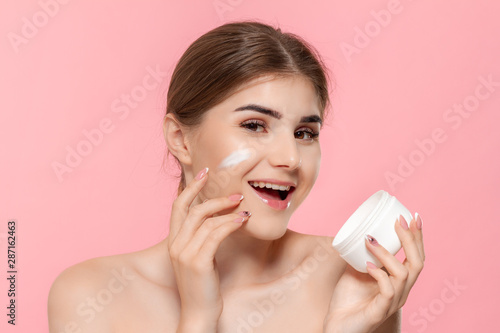 Beautiful girl applying moisturizing cream from tube to skin. Model looks at the camera isolated over pink background. Concept of beauty and health treatment.