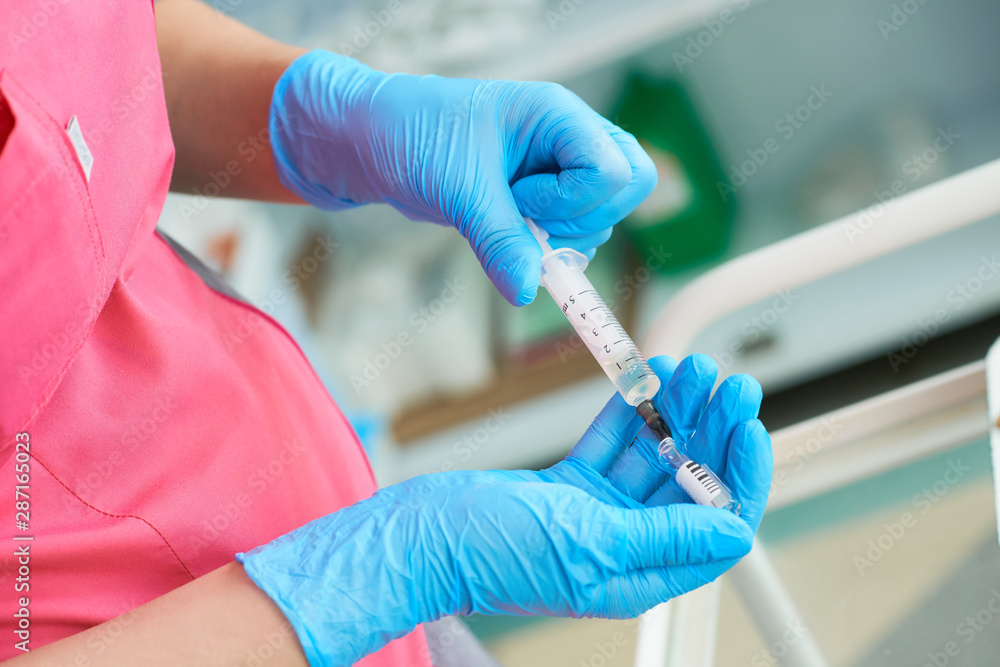 Close-up hands of young woman doctor anesthesiologist dressed in pink gown, blue gloves prepares solution for anesthesia in the operating room before the surgery. Close-up hands of young woman doctor anesthesiologist dressed in pink gown, blue gloves prepares solution for anesthesia in the operating room before the surgery.