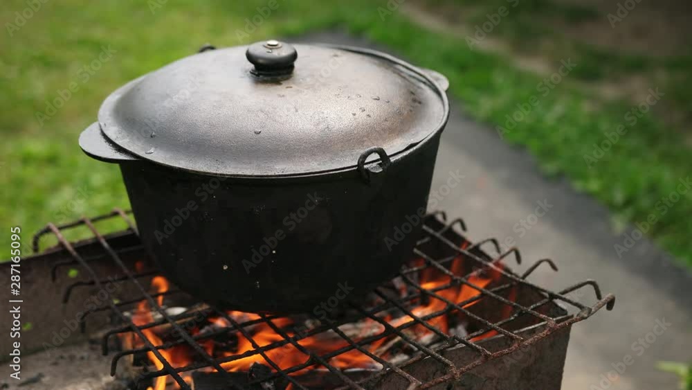 picnic in nature, fire burns on the grill and dish is cooked in large cast-iron cauldron, covered with a lid, in the day, around the beautiful green grass, close-up, slow motion picnic in nature, fire burns on the grill and dish is cooked in large cast-iron cauldron, covered with a lid, in the day, around the beautiful green grass, close-up, slow motion