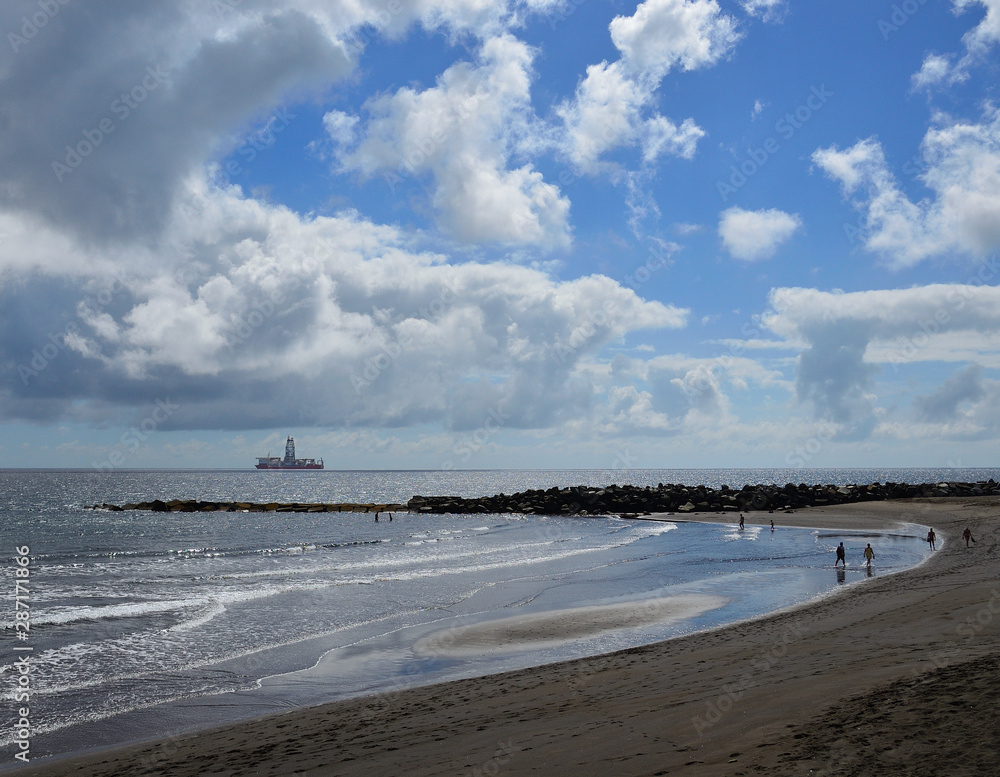 Sandy beach with people walking on the shore, blue sky with clouds and ship, La laja, coast of Las Palmas de Gran Canaria 