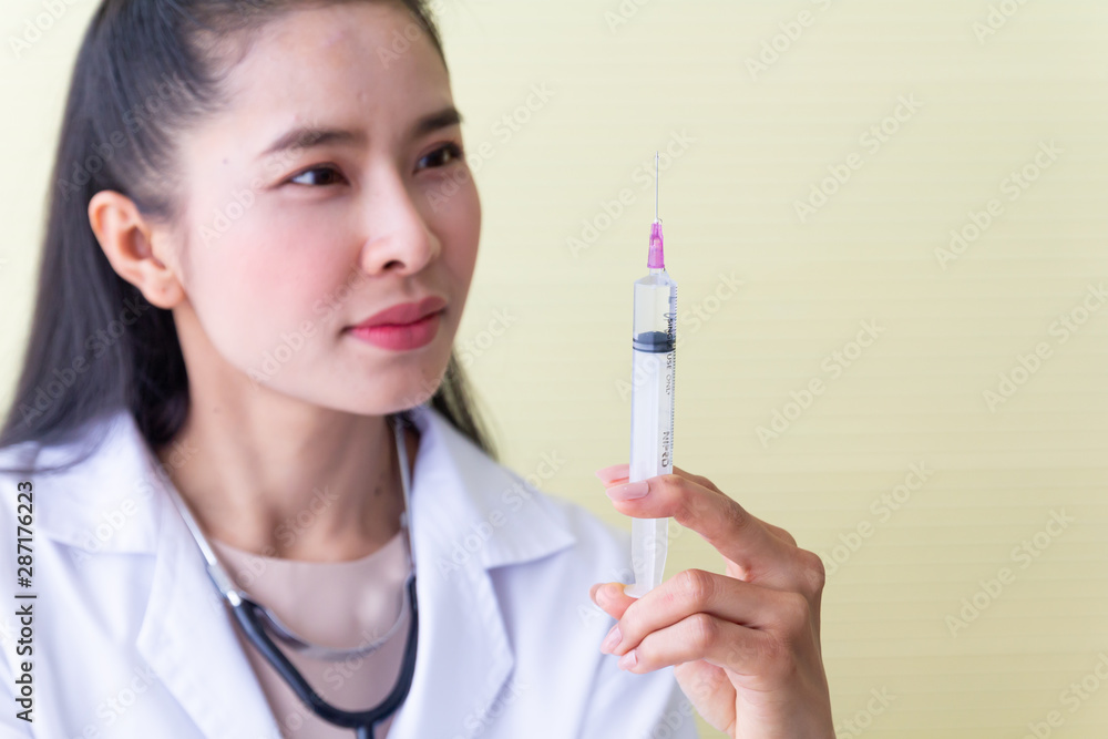 The female Asian doctor wearing a white robe sitting in a chair with a stethoscope on the neck be a serious face and preparing a syringe for patients. The female Asian doctor wearing a white robe sitting in a chair with a stethoscope on the neck be a serious face and preparing a syringe for patients.