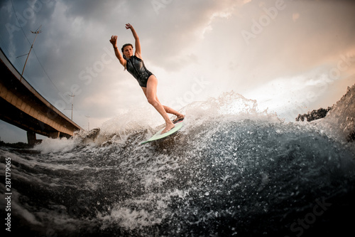 Sporty girl riding on the wakeboard on the river in the background of the bridge rising hands