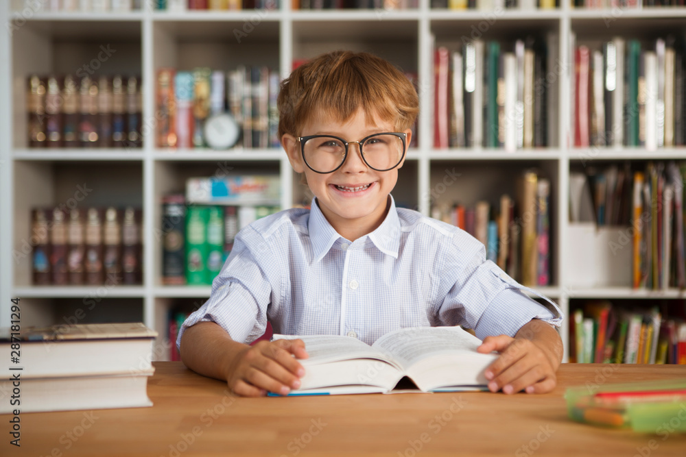 little boy wearing glasses reading book at table in library little boy wearing glasses reading book at table in library