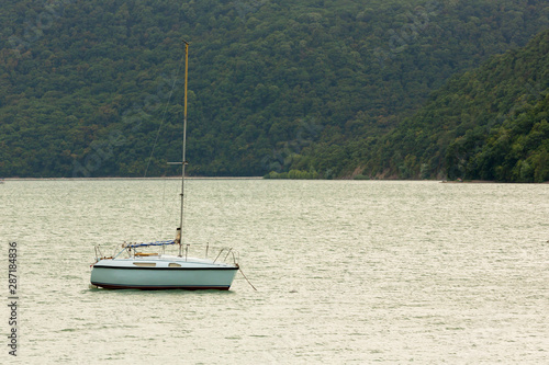 A sailing yacht is anchored on a lake in the background of a hill with a forest.