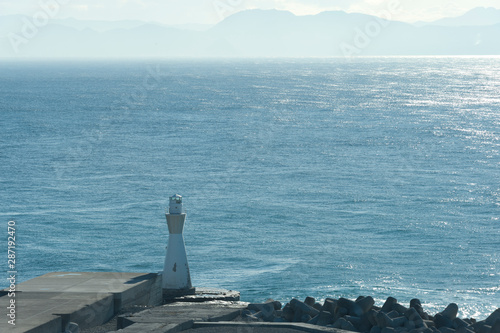 Blue sea with white lighthouse next to breakwater. Tagonoura Port - Fuji City, Japan. Background with mountains in fog.